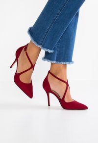 Kvinna i röda spetsiga högklackade skor med korsade ankelremmar och utsvängda blå jeans med fransade kanter mot en vit bakgrund.