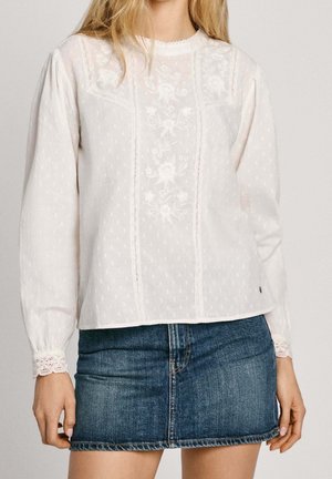 Femme portant un chemisier en dentelle blanc à manches longues avec broderies florales et une mini-jupe en denim bleu sur fond uni.