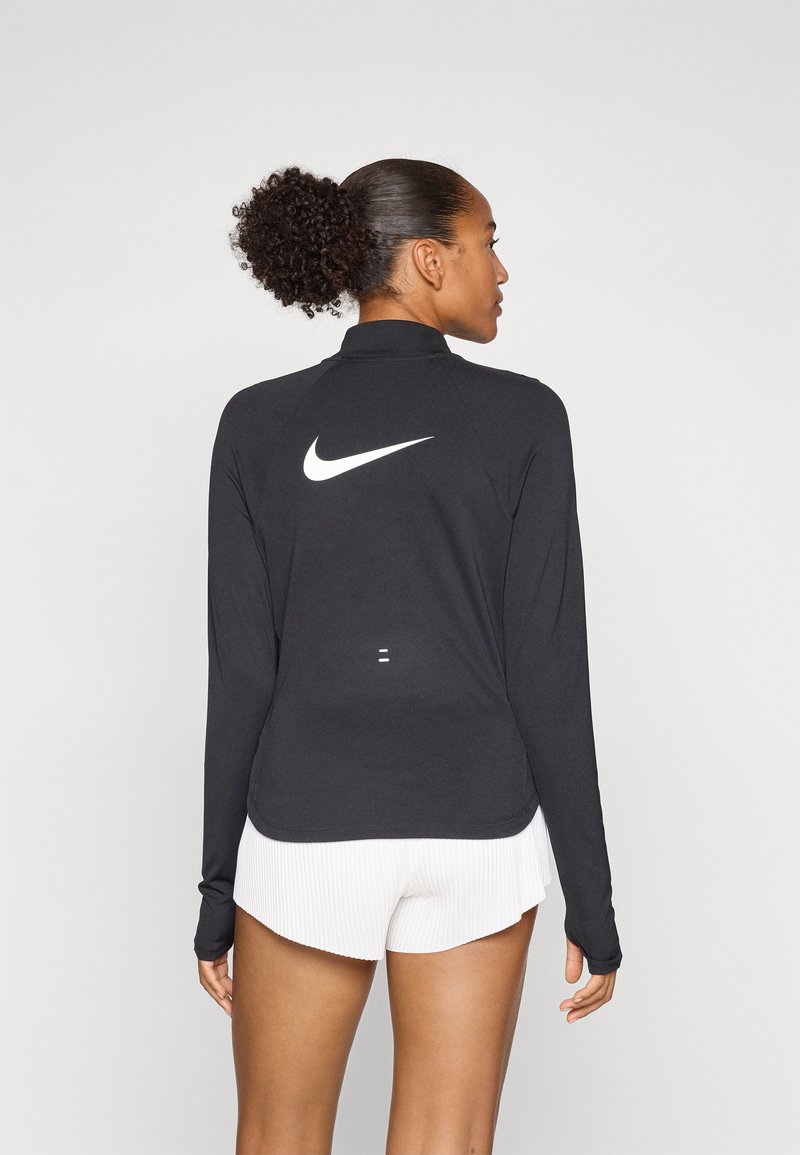 Nike black long-sleeve top with a high collar, featuring a white logo on the back, paired with white pleated shorts. Smooth, lightweight fabric.