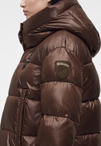Vue de côté d'une personne portant une doudoune marron brillante avec une grande capuche et un patch logo Blauer sur la partie supérieure de la manche.