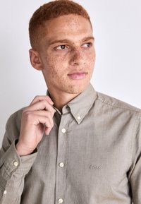 Camisa de botones verde claro con cuello, que presenta un logo en el pecho, botones blancos y una tela texturada. El modelo ajusta el cuello.