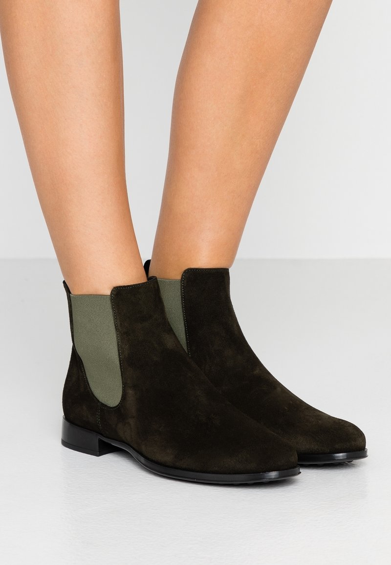 Bottes Chelsea en daim noir avec des panneaux latéraux élastiques vert foncé, portées sur des jambes nues sur un fond clair uni.