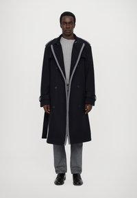 Cappotto doppiopetto blu navy con accenti grigi, dotato di ampie rever, tasche laterali e cintura. Abbinato a un maglione grigio a coste e jeans grigi.