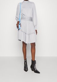 Robe grise à manches longues avec une taille cintrée, une jupe évasée et un ourlet à volants. Associée à des bottines noires et un petit sac bandoulière bleu.