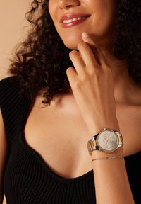 Silber- und goldfarbene Edelstahl-Uhr mit rundem Ziffernblatt, Kristallakzenten und drei Hilfszifferblättern an einem Handgelenk neben einem zarten Armband.