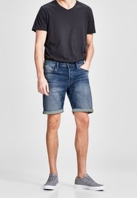 Muž v černém tričku s krátkým rukávem, s ohrnutými modrými denimovými šortkami a šedými teniskami, stojící s jednou rukou v kapse na bílém pozadí.