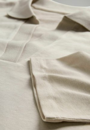 Tricou polo bej din bumbac, cu guler clasic, mâneci pliate și detalii vizibile de cusătură. Textura pare netedă și ușor texturată.