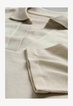 Beiges Baumwoll-Poloshirt mit klassischem Kragen, umgeschlagenen Ärmeln und sichtbaren Steppnähten. Die Textur wirkt glatt und leicht strukturiert.