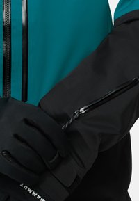 Wasserfeste Jacke in Teal und Schwarz mit einer Reißverschlusstasche am Arm, ausgestattet mit einem engen Bündchen und detailliertem strukturiertem Stoff.