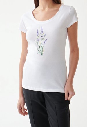 Kobieta w białej koszulce z krótkim rękawem z motywem lawendy i czarnych spodniach, stojąca na jednolitym białym tle.