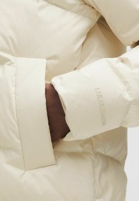 Creme pufferjakke med quiltet tekstur, med en lomme og præget logo på ærmet. Letvægts, glat stof.