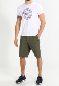 Camiseta blanca de algodón con un logo gráfico en rojo y azul marino, combinada con pantalones cortos verde oliva. Lleva zapatillas azul marino y el modelo se encuentra en un fondo neutro.