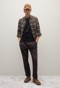 Braunes kariertes Hemd mit Knöpfen über einem schwarzen T-Shirt, kombiniert mit dunklen Hosen und braunen Wildlederschuhen. Eine beige Baseballkappe mit "P"-Logo.