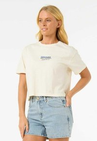 Skórzana koszulka w kolorze kremowym z krótkimi rękawami, z niebieskim nadrukiem logo "Rip Curl Surfing Co" z przodu, w zestawie z jasnoniebieskimi szortami dżinsowymi.