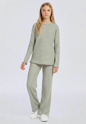 Jonge persoon staat en draagt een lichtgrijs en wit gestreept lange mouwen top en broek met golvende randen en witte sneakers tegen een effen achtergrond.