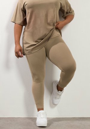 Kvinde iført beige oversized t-shirt og leggings med hvide sneakers, stående mod en hvid væg med det ene knæ bøjet.