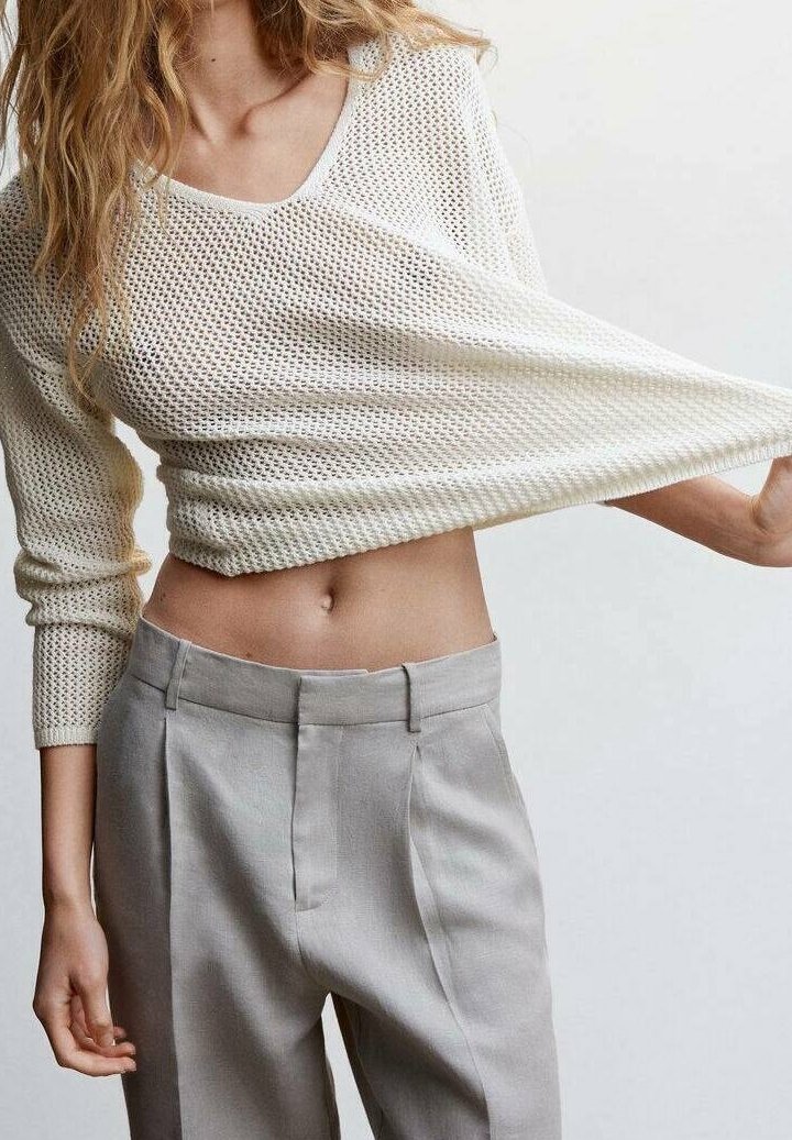 Weißer, kurzer Strickpullover mit V-Ausschnitt und langen Ärmeln, kombiniert mit hellgrauen, hoch taillierten Hosen mit Frontfalten.
