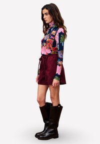 Frau steht seitlich mit einem bunten Rollkragenpullover, einem weinroten Wickelrock und schwarzen Kniestrümpfen vor einem weißen Hintergrund.