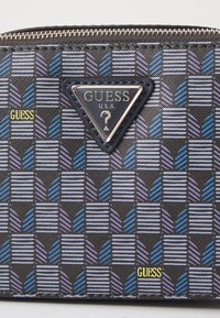 Portafoglio nero con motivi geometrici blu e viola, caratterizzato da un prominente logo triangolare in argento. Il testo "GUESS" è incorporato in giallo.