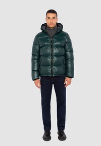 Grøn puffet jakke med glinsende finish, lynlås foran og hætte. Båret over en grå sweater, kombineret med mørke jeans og sorte sko.