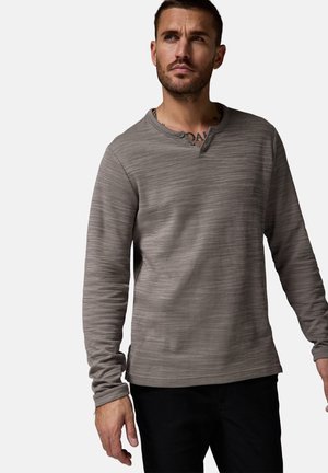 Langärmliges, graues Henley-Shirt mit einem strukturierten Muster, rundem Ausschnitt und drei Knöpfen am Kragen. Dunkle Hose wird getragen.