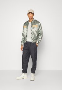 Satin olive-green bomber jacket with orange lettering, beige cap, gray shirt, dark trousers, and white lace-up boots.