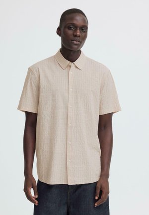 Homme portant une chemise beige à manches courtes et à boutons, debout devant un fond blanc uni.