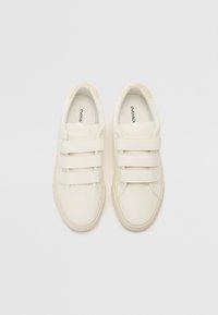 Baskets blanches avec trois bandes Velcro, en matériau synthétique lisse, forme arrondie et semelle en caoutchouc texturé. Accents de couleur crème au niveau du talon.