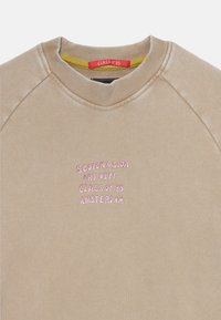 Béžová mikina s ribovaným výstrihom. Na prednej strane je ružový vyšívaný nápis: "SCOTCH & SODA ART DEPT. CLASS OF '85 AMSTERDAM." Hladká textúra.