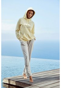 Geel hoodie met een voorzak en bedrukte tekst; gecombineerd met lichtgrijze sweatpants. Model staat blootvoets bij water.