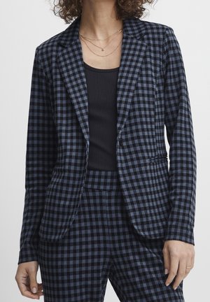 Mujer vistiendo una chaqueta de cuadros en negro y azul y pantalones a juego con un top negro acanalado y collares dorados en capas.