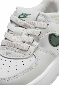 Nahaufnahme eines weißen Nike-Sneakers mit strukturierten Schnürsenkeln, perforierter Zehenpartie, grünem Logo auf der Zunge und der Seite sowie detaillierter Nahtführung.