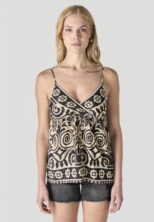 Mujer rubia con top sin mangas de tirantes finos, estampado en negro y beige, y pantalones cortos de mezclilla negra, de pie frente a un fondo gris.