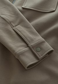Beige fabric shirt with a textured finish, featuring a rolled sleeve with a button closure and a slight sheen from the material.