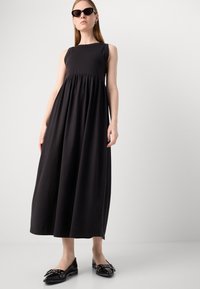 Svart langermet maxikjole med rynket liv, laget av myk stoff. Kombinert med skinnende svarte flate sko med sløyfe detaljer. Elegant helhetsdesign.