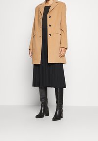 Beige Wollmantel mit Reverskragen, drei Knöpfen und zwei Taschen, getragen über einem schwarzen plissierten Kleid und kombiniert mit knielangen schwarzen Stiefeln.