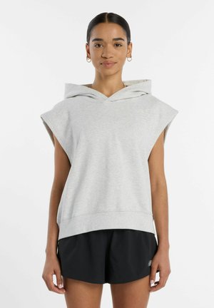 Kvinde med mørkt hår iført lysegrå ærmeløs hættetrøje og sorte shorts, står foran hvid baggrund, vendt mod fronten.