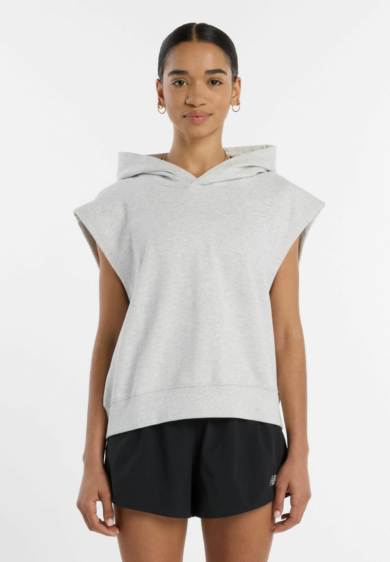 Femme aux cheveux foncés portant un sweat à capuche sans manches gris clair et un short noir, debout devant un fond blanc, face à l'avant.