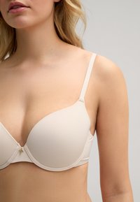 Beige gevoerde beha met verstelbare banden, voorzien van een soepele textuur en subtiele stiksels, afgewerkt met een klein gouden decoratief detail in het midden.