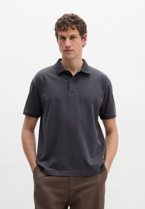 Man met krullend haar, gekleed in een donkergrijs poloshirt met korte mouwen en een bruine broek, staand met handen in de zakken tegen een effen achtergrond.