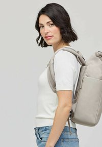 Femme aux cheveux foncés portant une chemise blanche et un jean bleu, portant un sac à dos gris clair sur l'épaule, devant un fond uni.