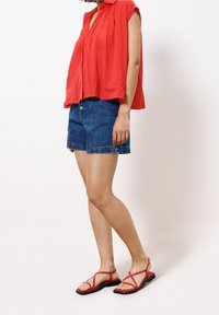 Persona vestida con una blusa roja sin mangas y holgada, pantalones cortos de mezclilla azules, y sandalias planas rojas con tiras, de pie contra un fondo blanco liso.