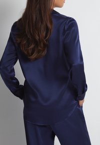 Blouse en satin bleu marine à manches longues, avec un ourlet légèrement arrondi et des poignets boutonnés, présentant une texture lisse et brillante.