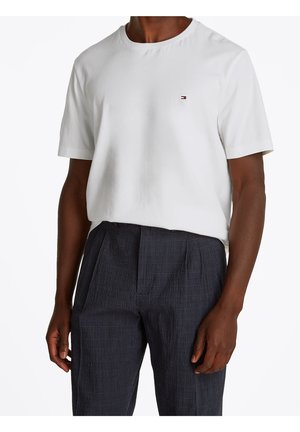 Weißes Baumwoll-T-Shirt mit rundem Ausschnitt und kurzen Ärmeln, kombiniert mit dunklen, strukturierten karierten Hosen. Rote und blaue Logodetails vorhanden.