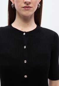 Μαύρο κοντομάνικο τοπ με ρίγες, με στρογγυλή λαιμόκοψη και πέντε διακοσμητικά κουμπιά με ασημένιες λεπτομέρειες κατά μήκος του εμπρός μέρους.