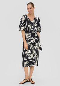 Robe portefeuille à motif noir et blanc avec des manches courtes, un décolleté en V et une ceinture assortie à la taille. Des sandales plates complètent le look.