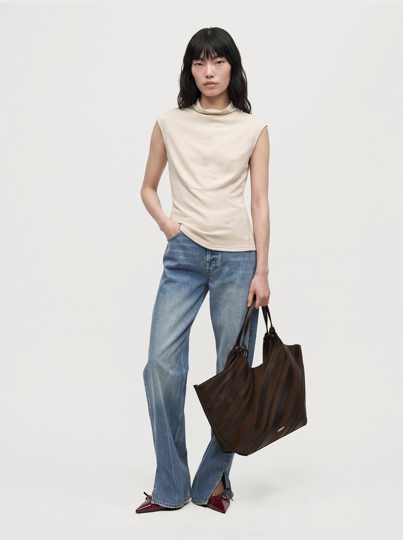 Un mannequin se tient devant un fond uni, vêtu d’un haut crème sans manches, d’un jean bleu et de talons bordeaux, et tient un grand cabas marron foncé.