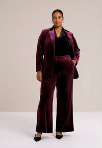 Lilla fløjlssuit med en tætsiddende blazer og vide bukser. Blazeren har en notch-lapel, båret over en top i sort blonde. Elegant silhuet.