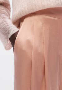 Pantalon plissé rose pâle en tissu lisse, avec une coupe ajustée et un léger éclat, associé à un pull rose clair doux et texturé.
