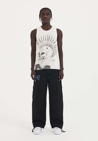 Weißes Tanktop mit grafischem schwarzen Totenkopf und Stacheln, kombiniert mit schwarzen Cargo-Shorts mit Taschen und blauen Logo-Details, getragen mit weißen Sneakers.